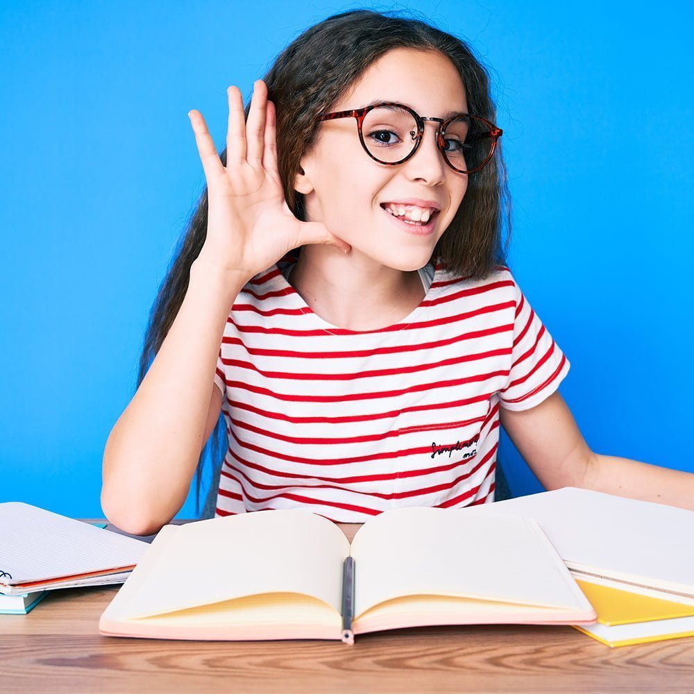 Young students with hearing loss sitting at desk with a notebook in front of her holding hand up to ear