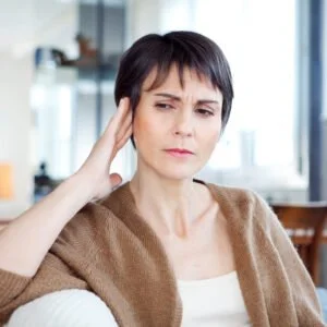 Middle-aged woman with short brown hair sitting down, lightly touching side of head with right hand with concerned look on face