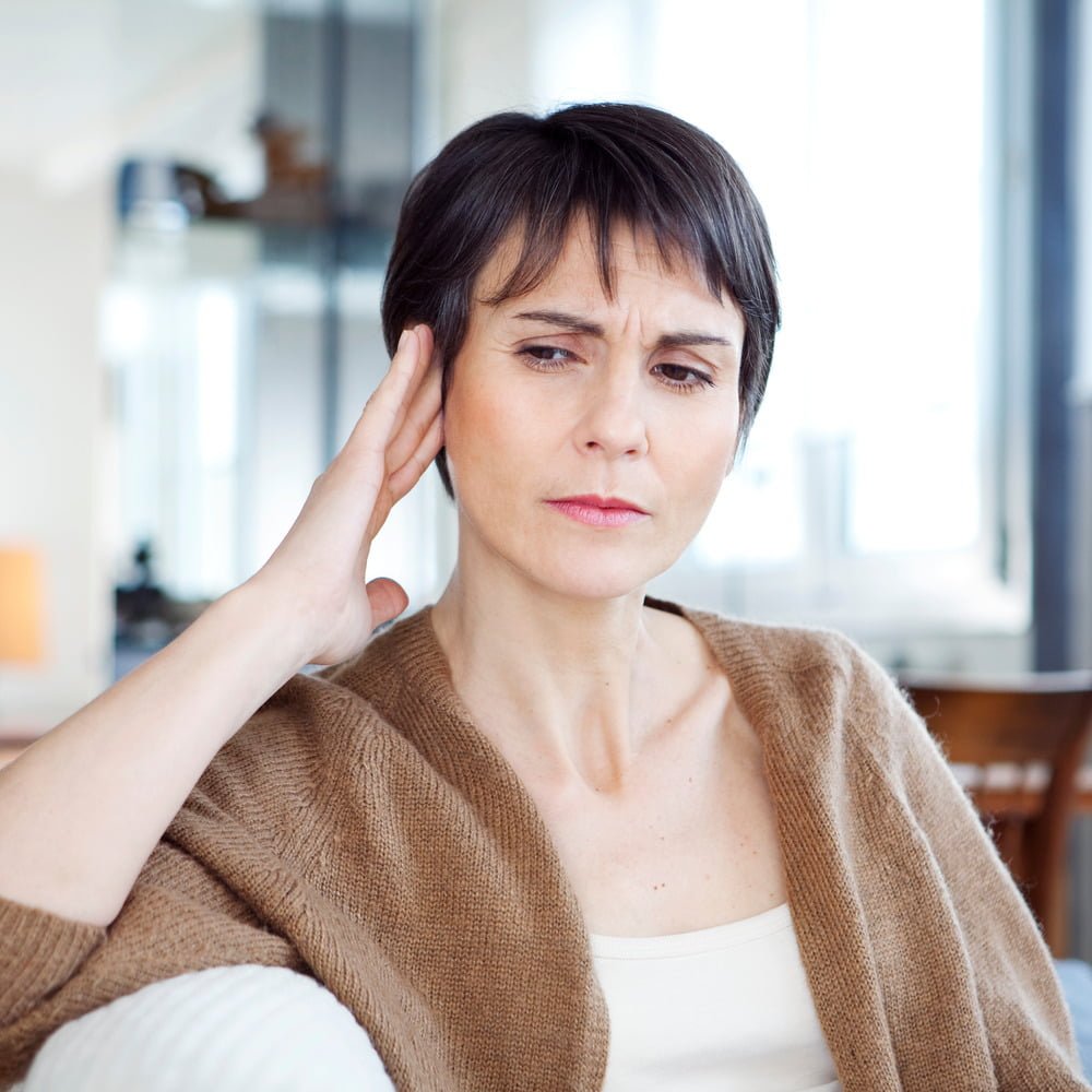 Middle-aged woman with short brown hair sitting down, lightly touching side of head with right hand with concerned look on face