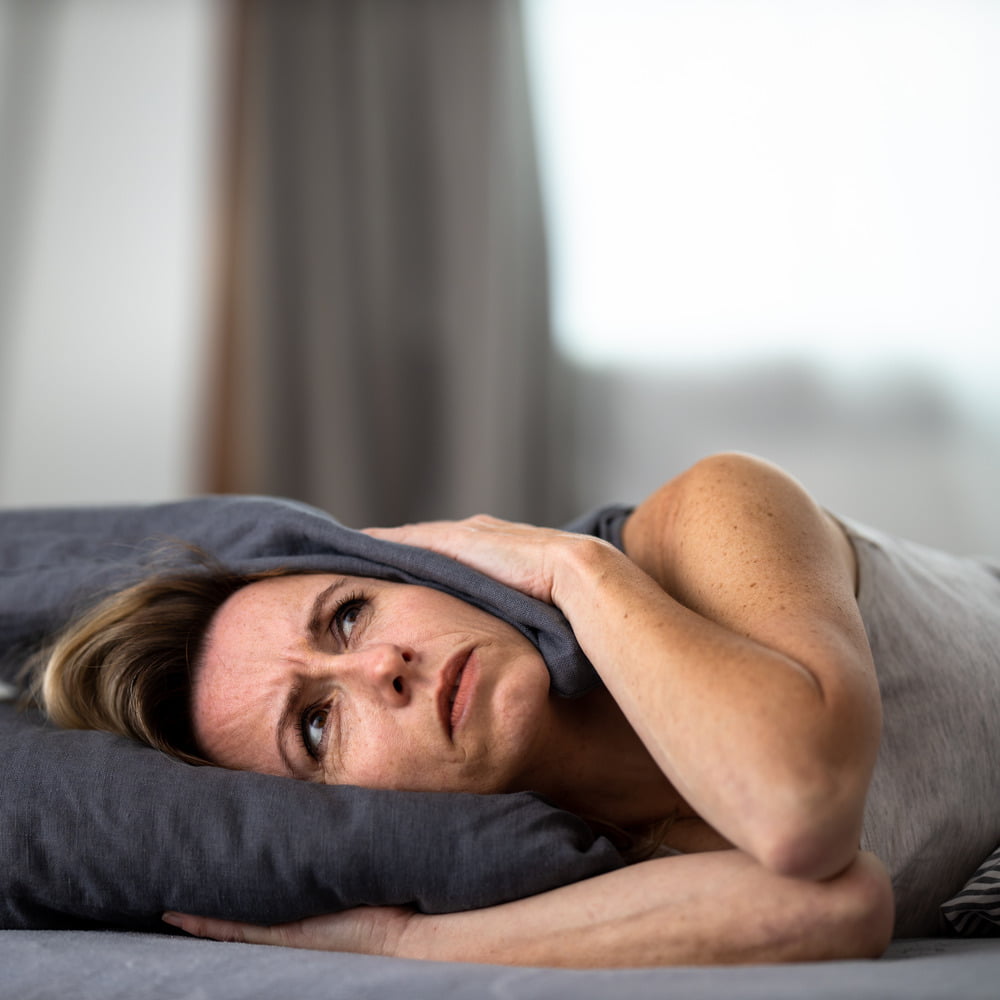 Middle-aged woman in bed, pressing both sides of pillow to head, visibly showing signs of tinnitus flare-up.