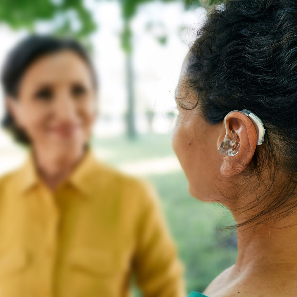 Close-up of a woman wearing a behind-the-ear hearing aid while conversing in a public places, highlighting the importance of hearing in social settings.