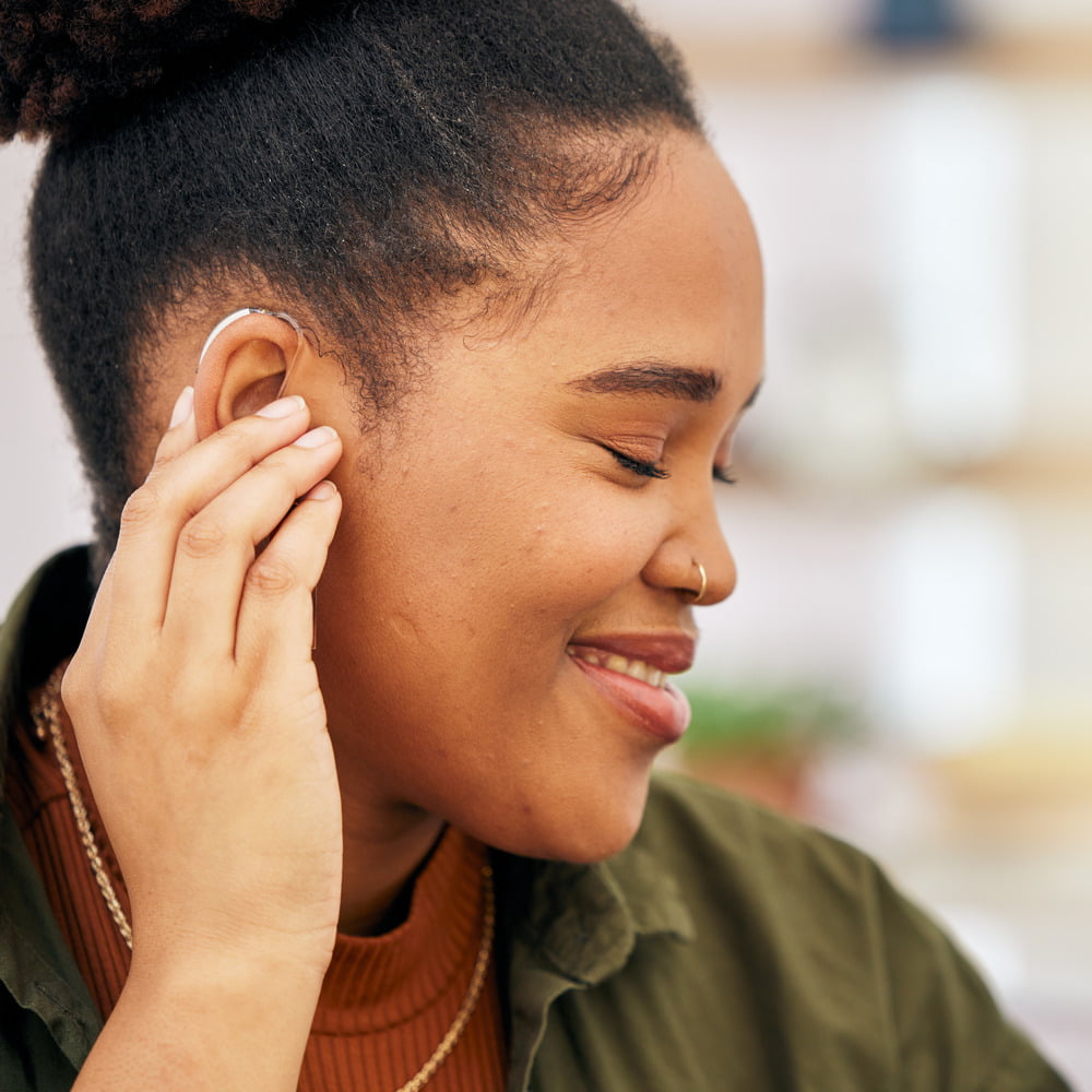Woman inserting a hearing aid into her right ear, looking down and smiling, promoting hearing health